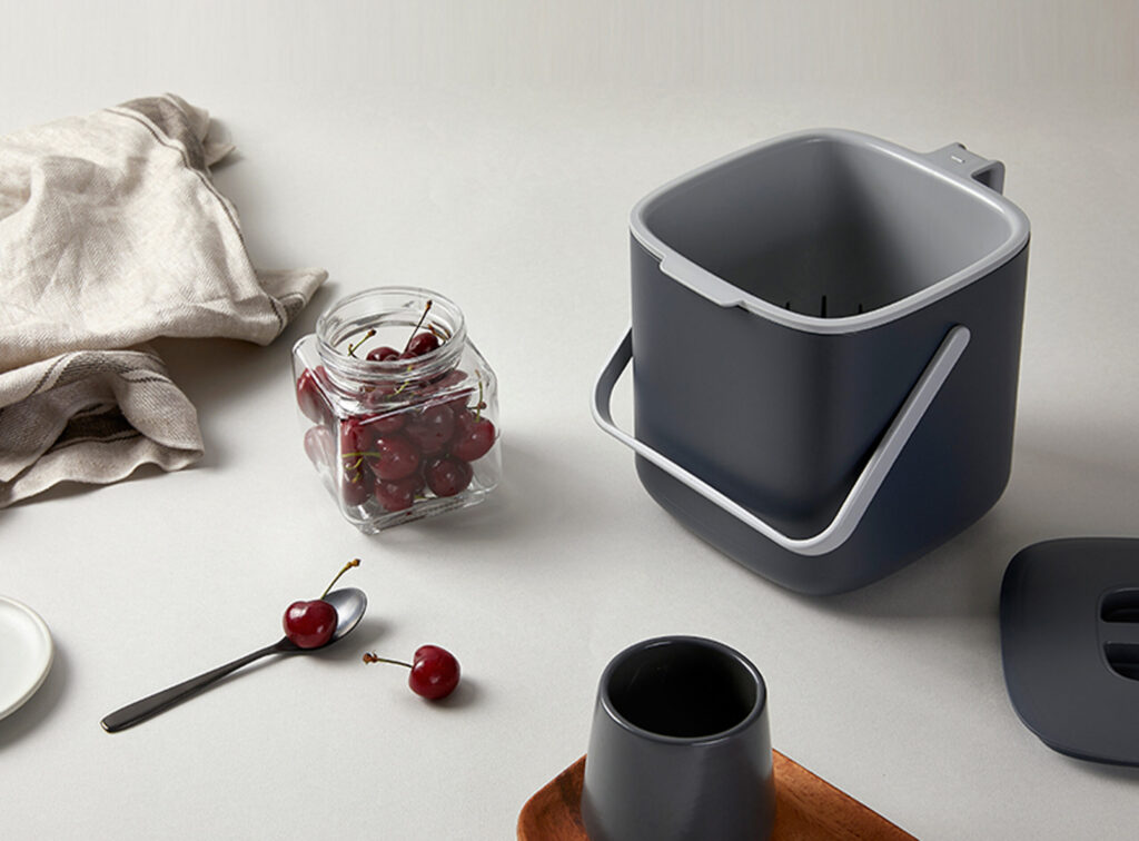 A premium kitchen lifestyle photograph of a charcoal-grey Maadan Food Composting Bin