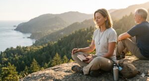 A man and woman meditating on a high mountain ridge overlooking a rugged coastline at golden hour, the woman holding a journal with her eyes closed, demonstrating peaceful mindfulness and relational harmony in nature.